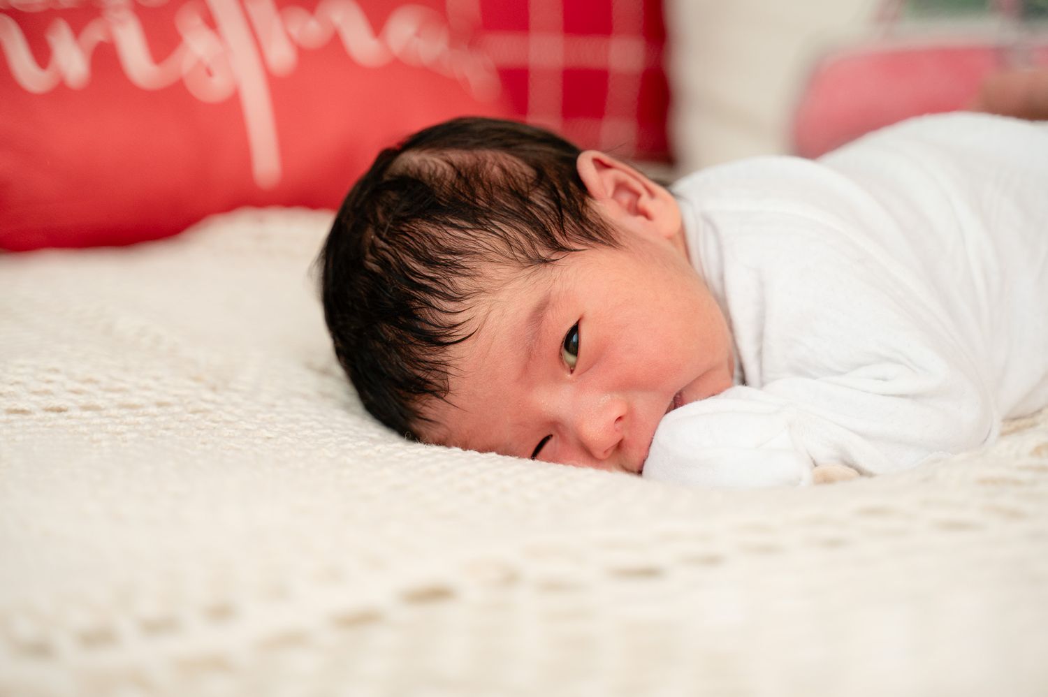 Newborn portrait on white bedding with red plaid pillow in background.