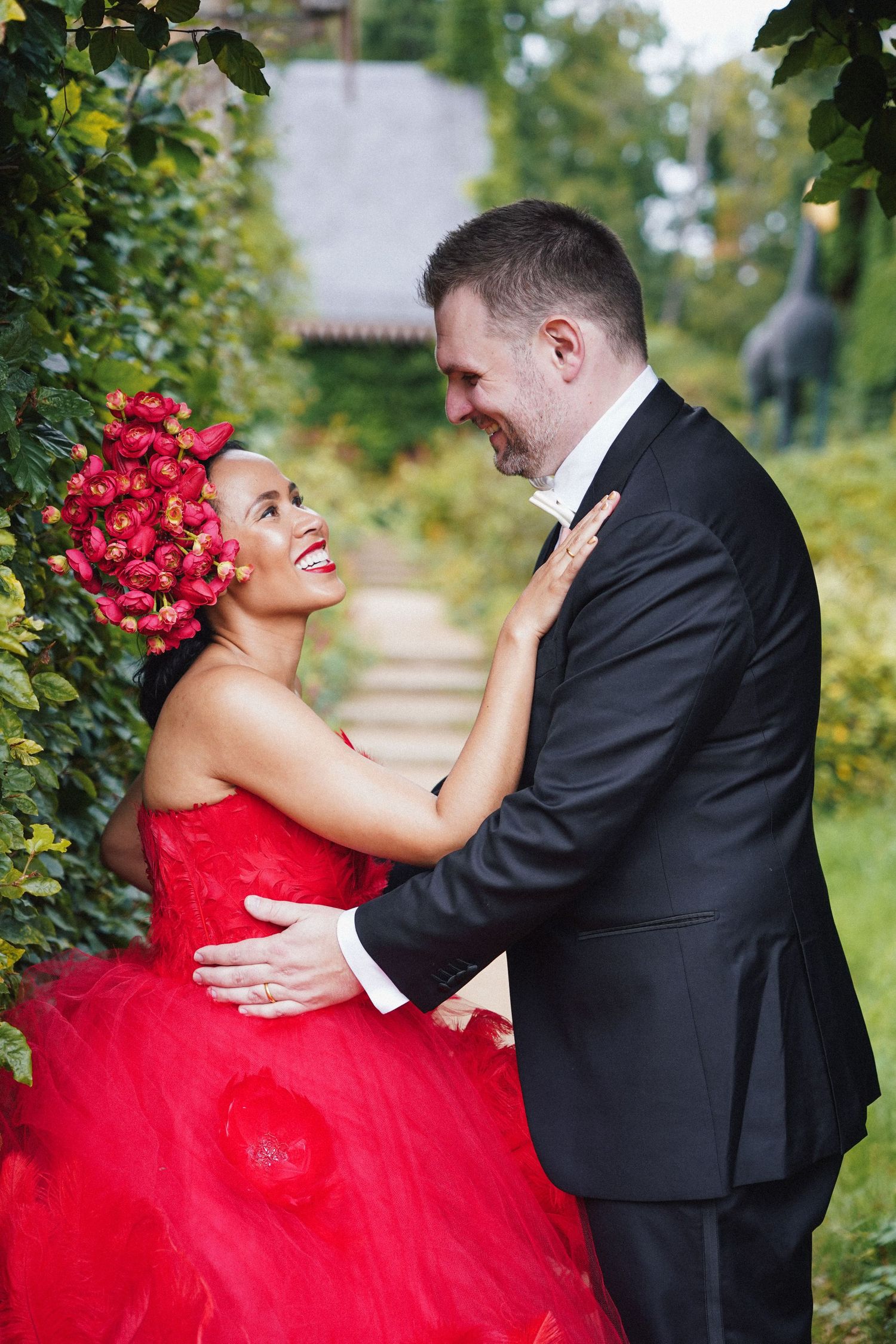 A couple in a romantic embrace outdoors, with the bride wearing a vibrant red ballgown and the groom in a black suit.