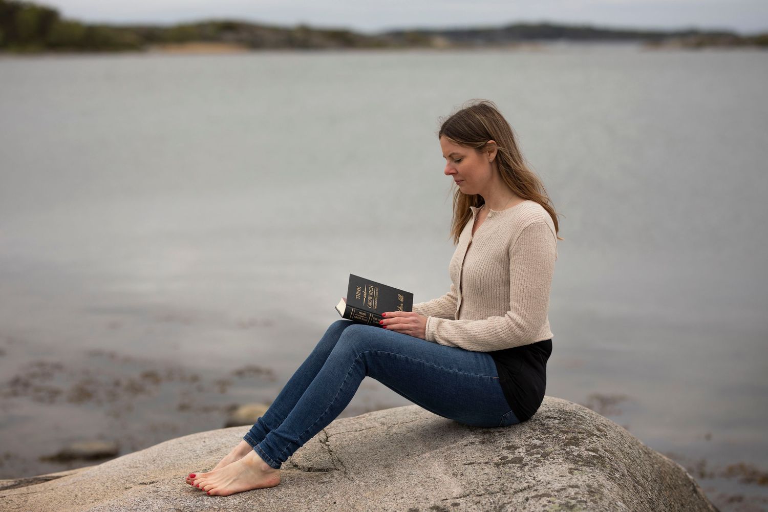 kvinna sitter på en klippa och läser en bok. i bakgrunden syns havet.