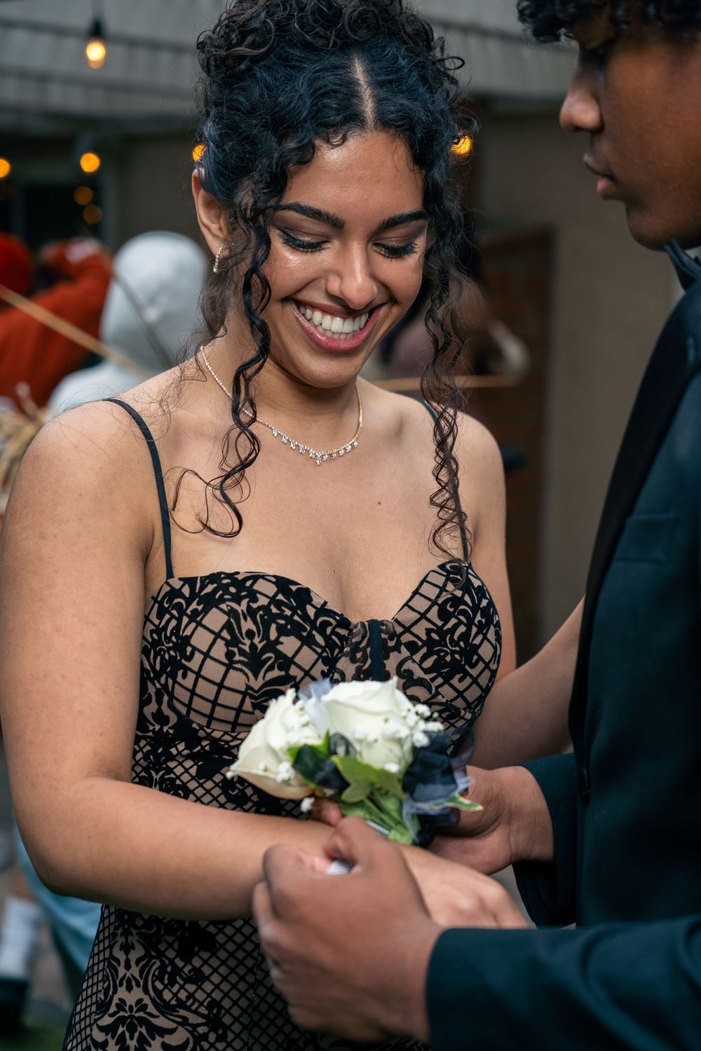Person in black lace dress smiles while holding a white flower corsage.