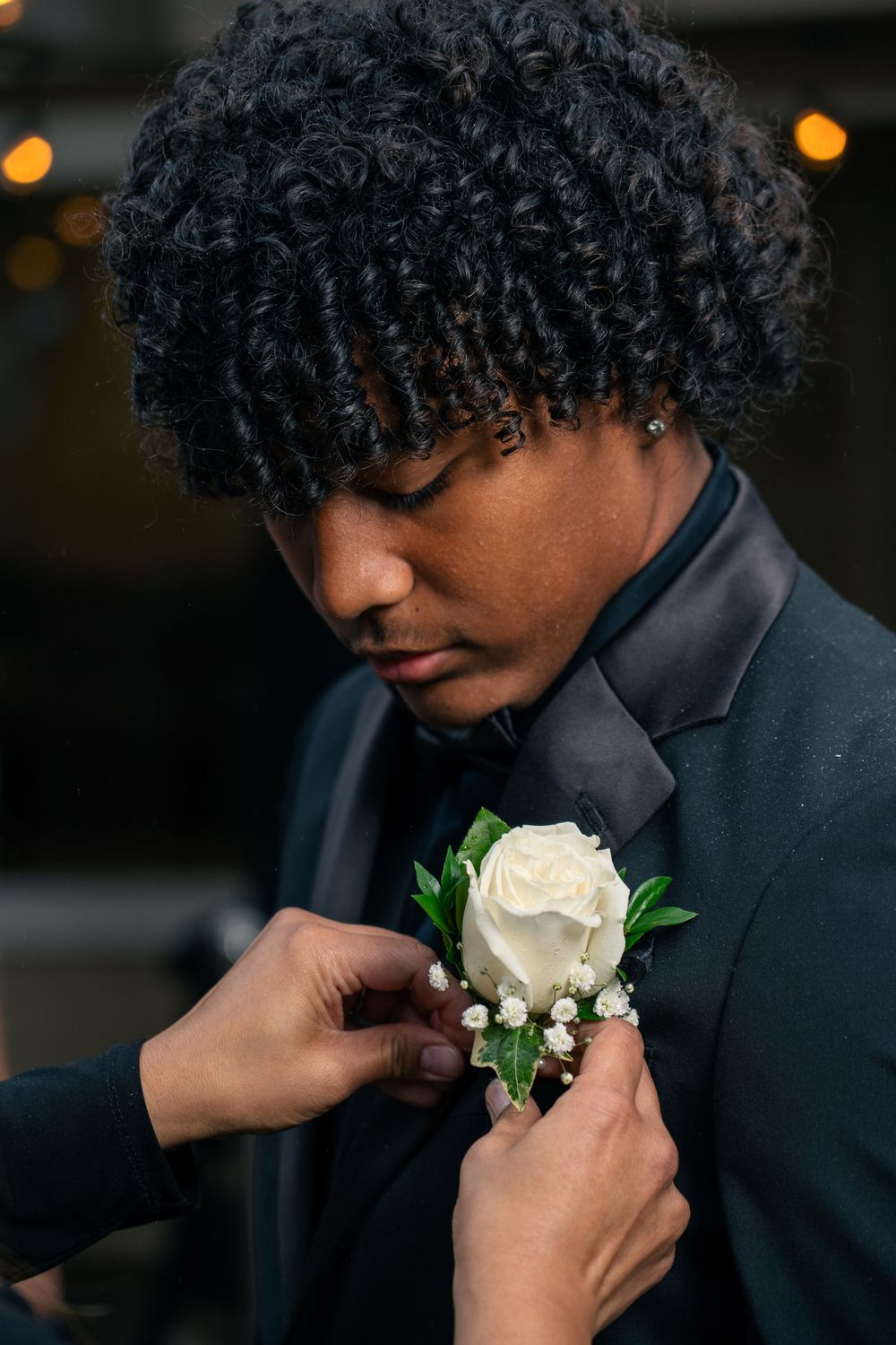 A side profile of someone getting a white rose boutonniere pinned to their black suit jacket.