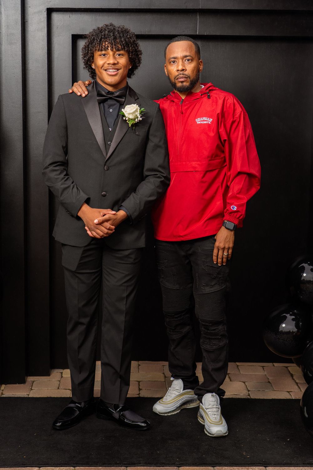 Two people pose together against dark wall, one in suit and one in red casual wear.