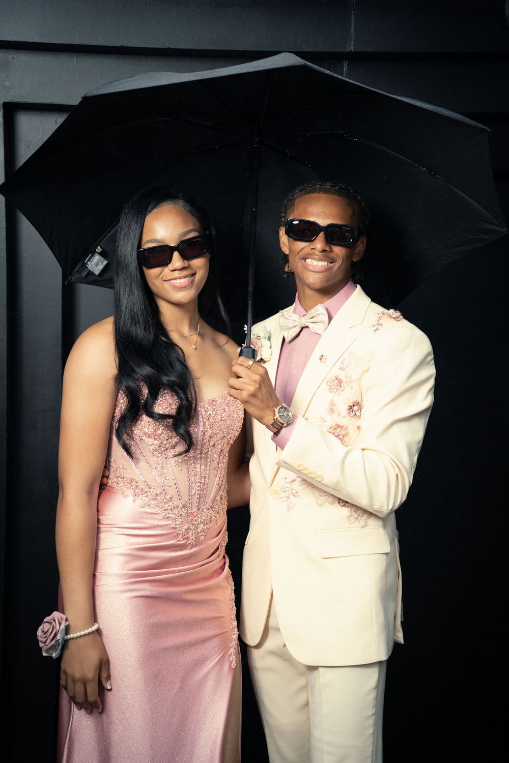 A couple in coordinated pink and cream formal wear poses with a black umbrella at an evening event.