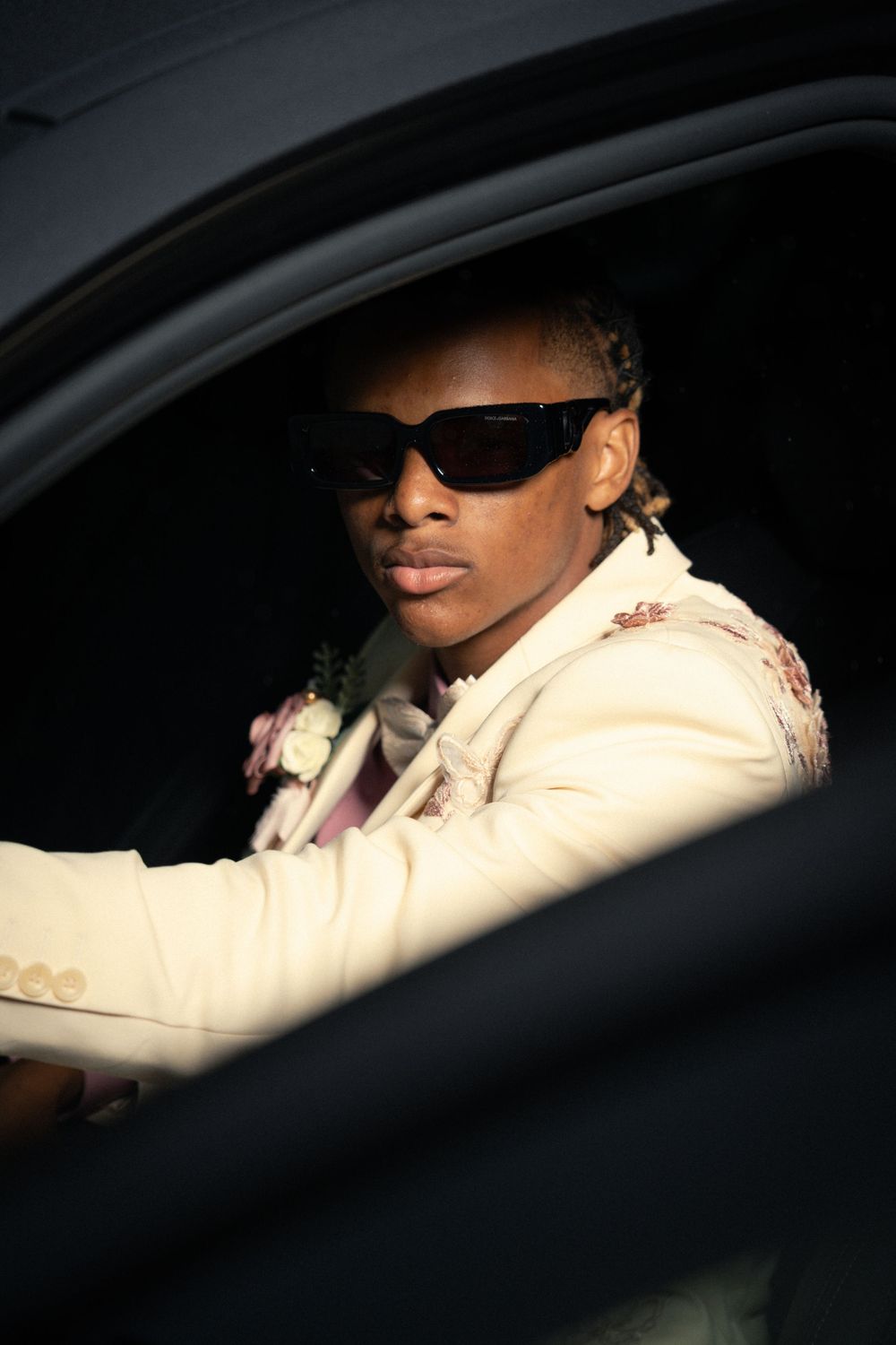 Person in cream colored formal wear and sunglasses seated in car.