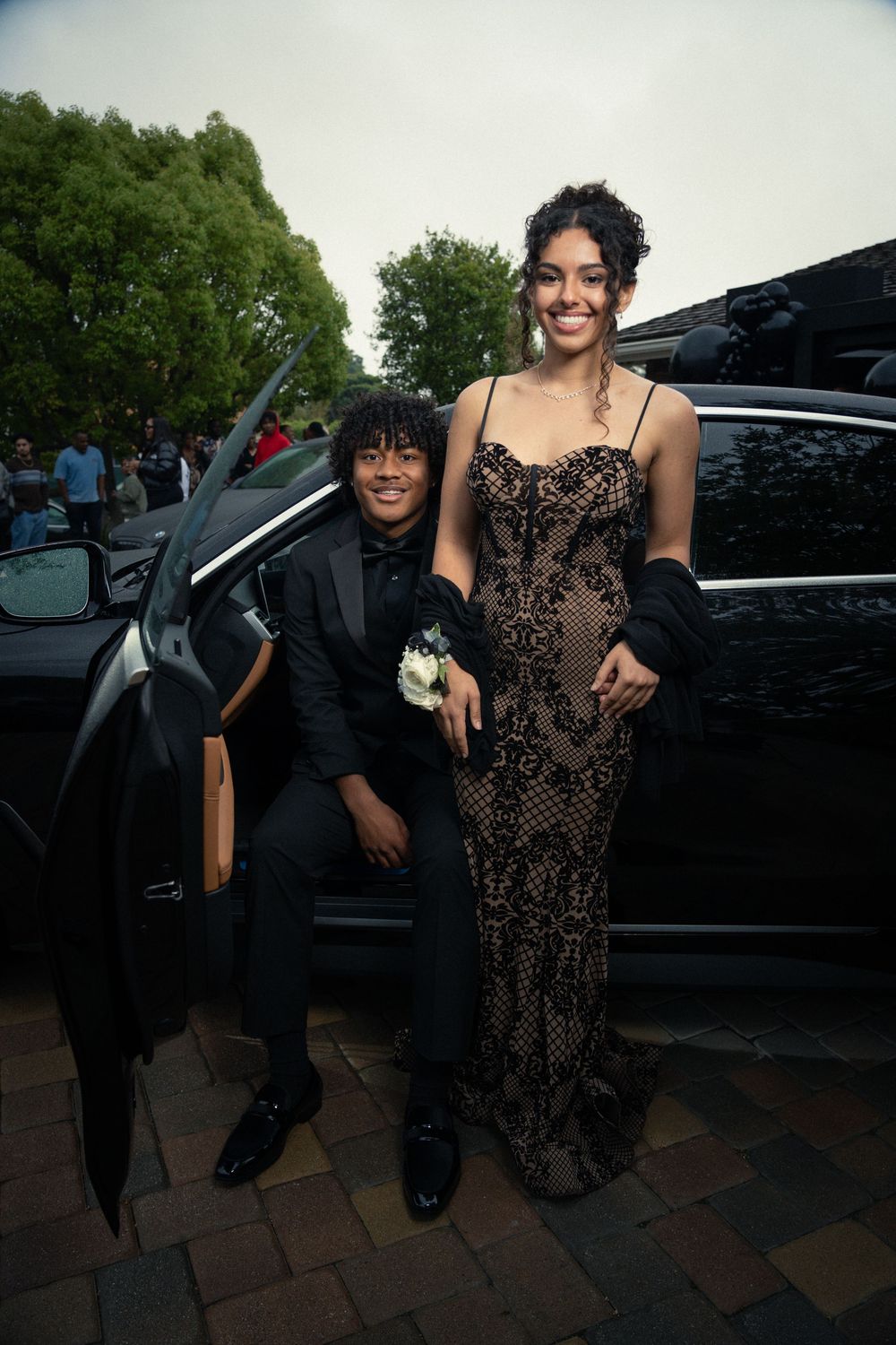 A pair in formal wear poses together by a black car, one in black and one in a lace patterned dress.