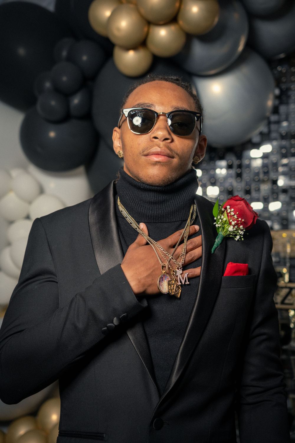 Person in black formal suit with turtleneck and red boutonniere poses against balloon backdrop.