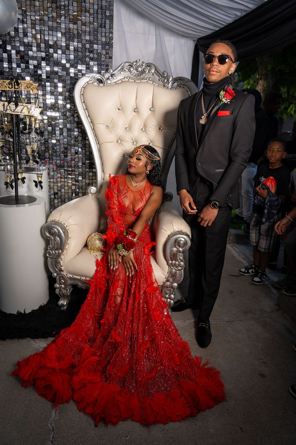 A couple in formal attire poses on an ornate white throne chair with a sparkly backdrop.