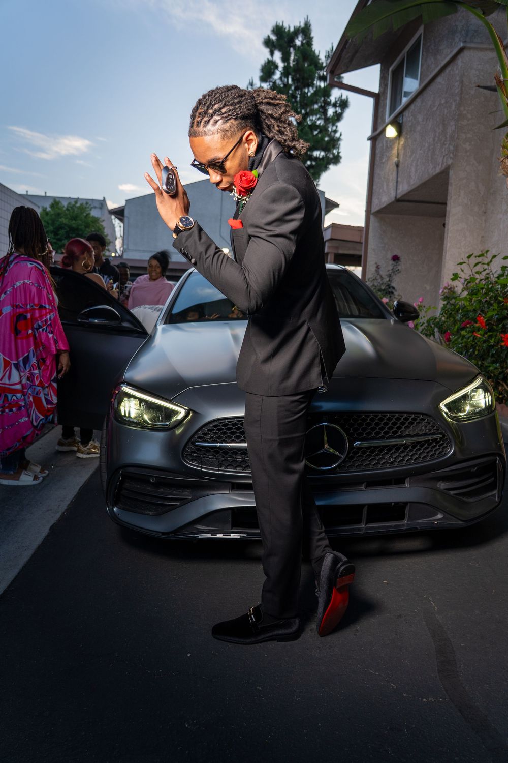 A person in a black suit poses dramatically in front of a Mercedes-Benz car at dusk.