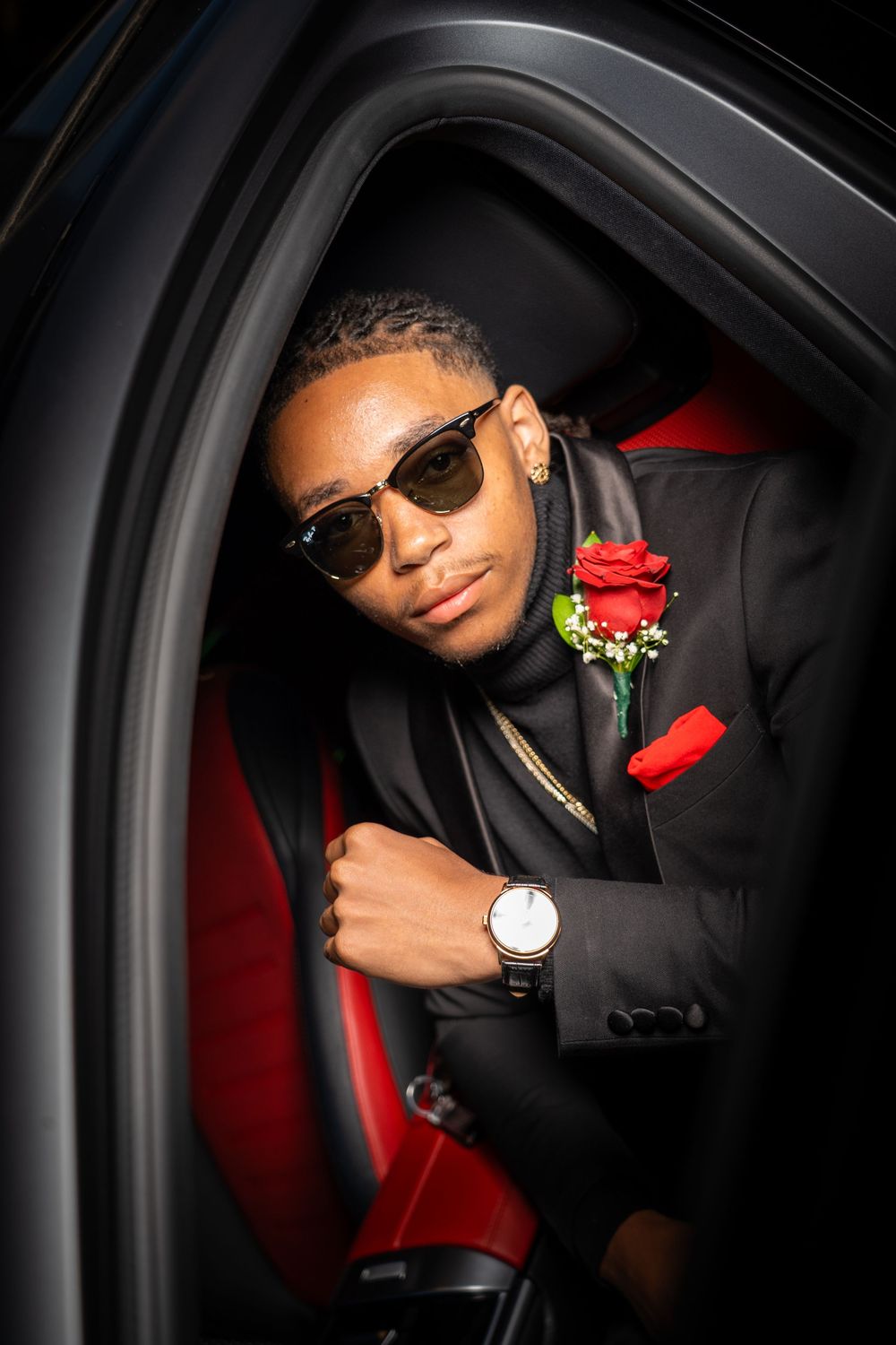 Person in black suit with red rose boutonniere poses in car with red interior.