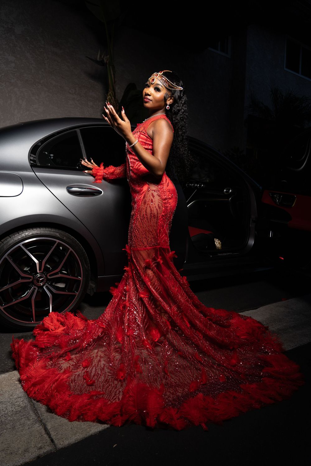 A dramatic photo of someone in a flowing red lace gown with a long train beside a luxury car.