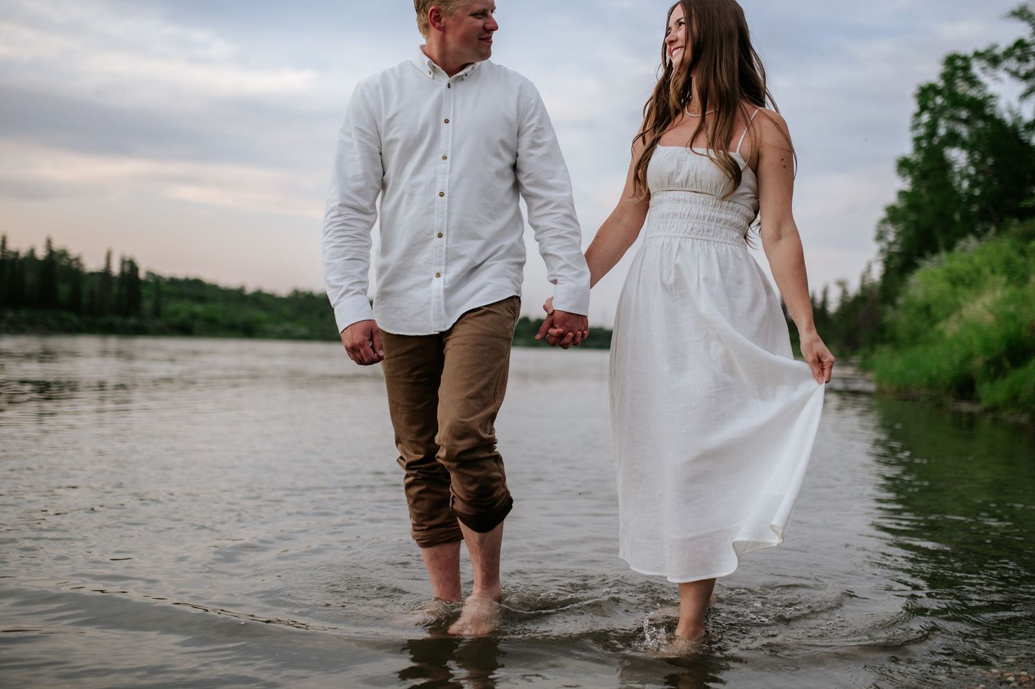 A couple holding hands while walking barefoot in shallow river water, wearing casual white wedding attire.