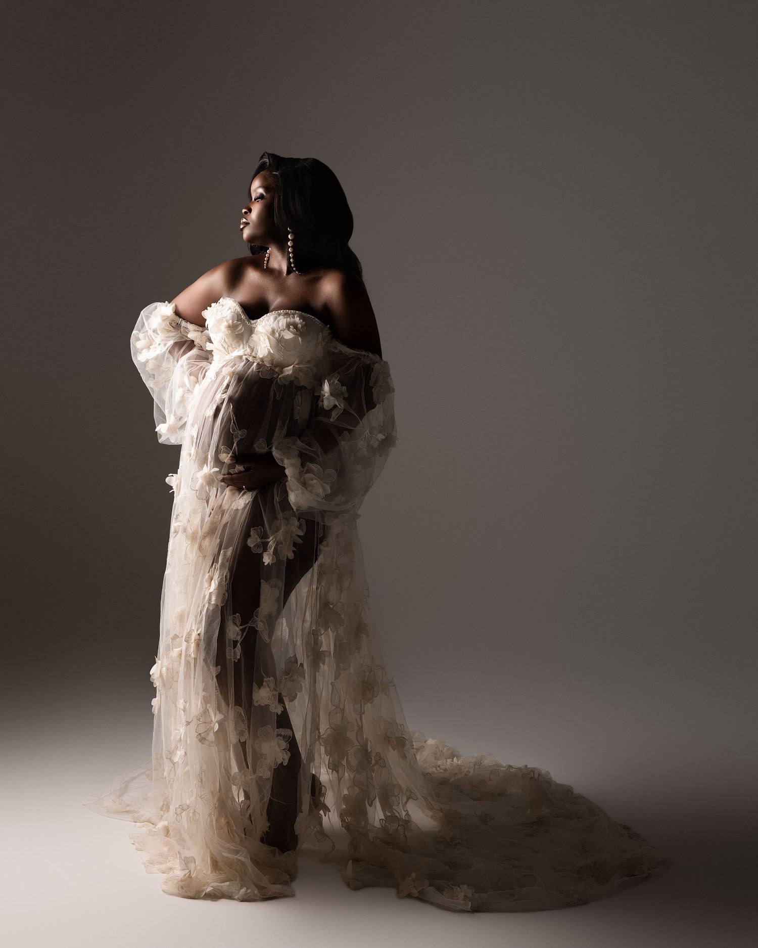 A Black woman poses elegantly in a sheer floral tulle gown with a dramatic train in a studio setting.