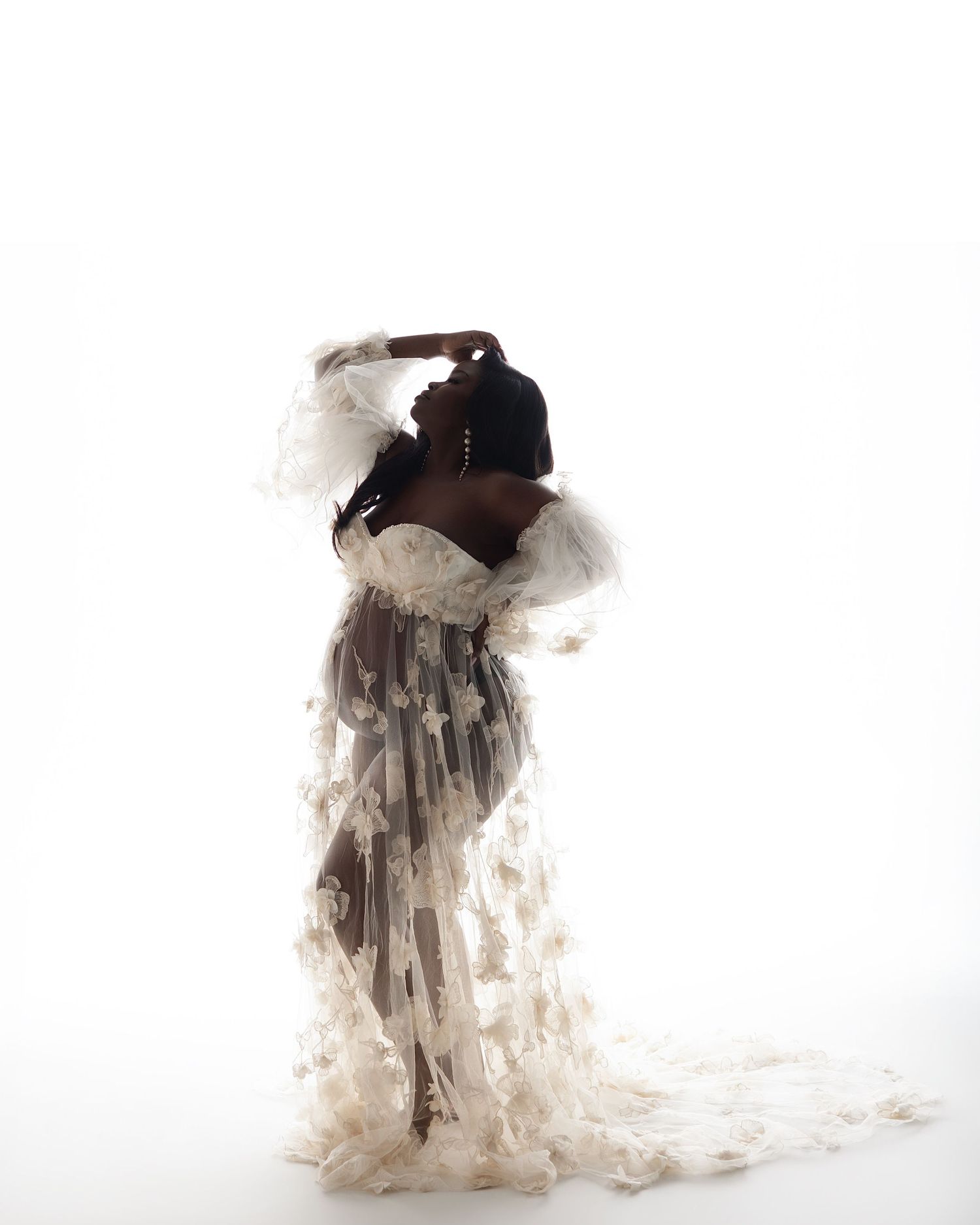 A Black woman poses gracefully in a sheer white floral gown with ruffled sleeves against a bright white background.