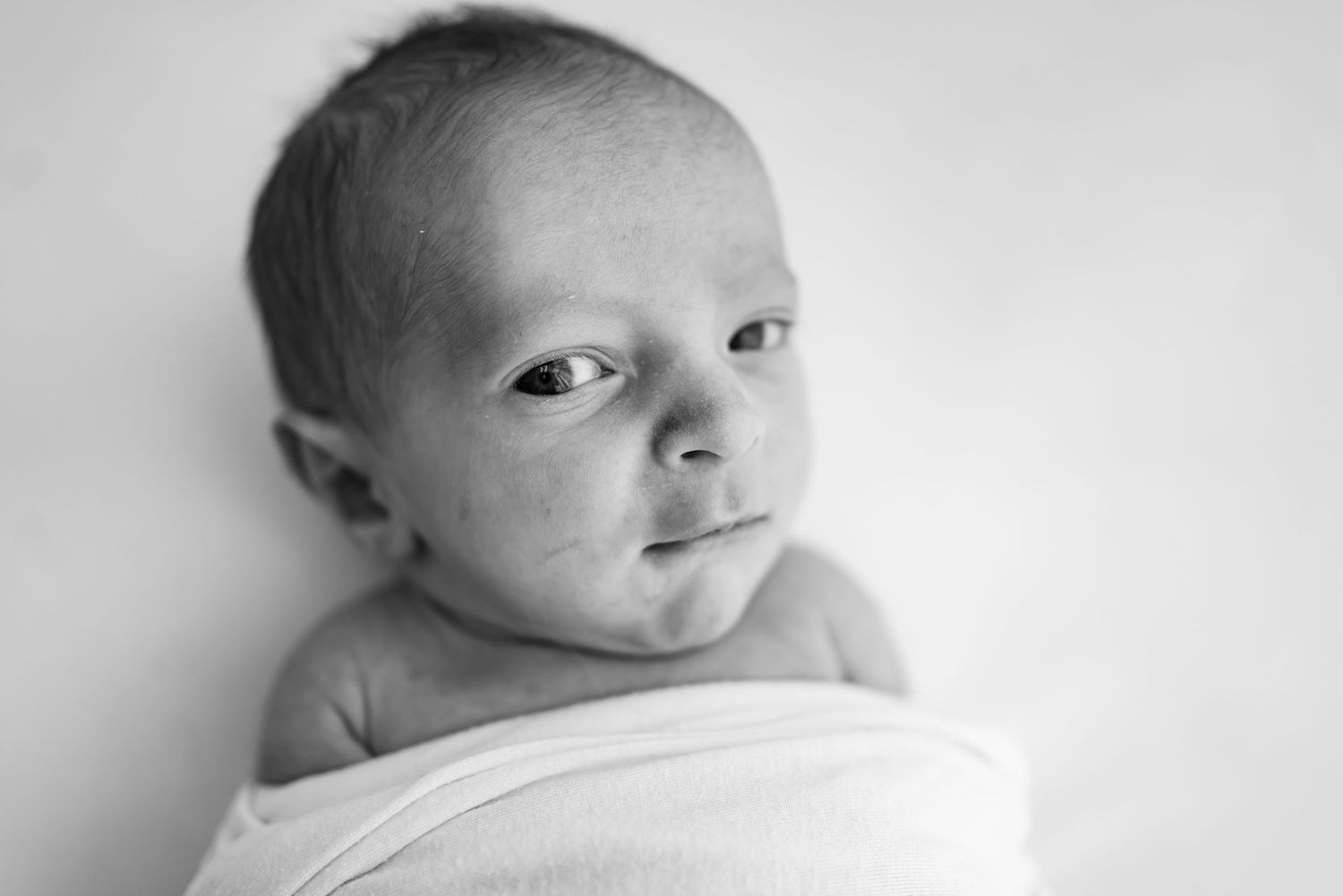 Black and white portrait of a tiny newborn baby wrapped in soft white fabric looking at camera.
