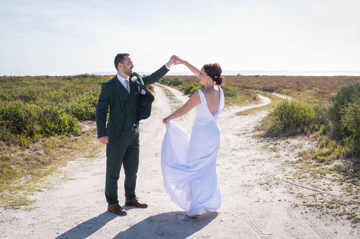 A couple in wedding attire walks down a sandy beach path bordered by green coastal vegetation.