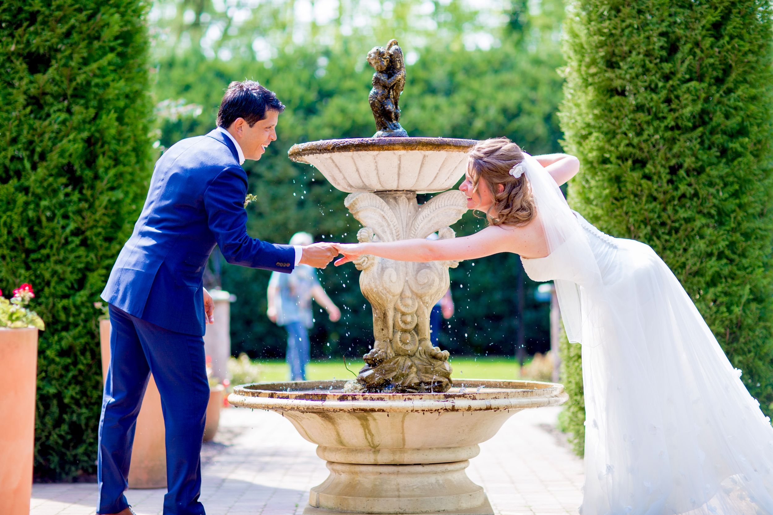 Newlyweds playfully pose by an ornate garden fountain during their wedding photoshoot.