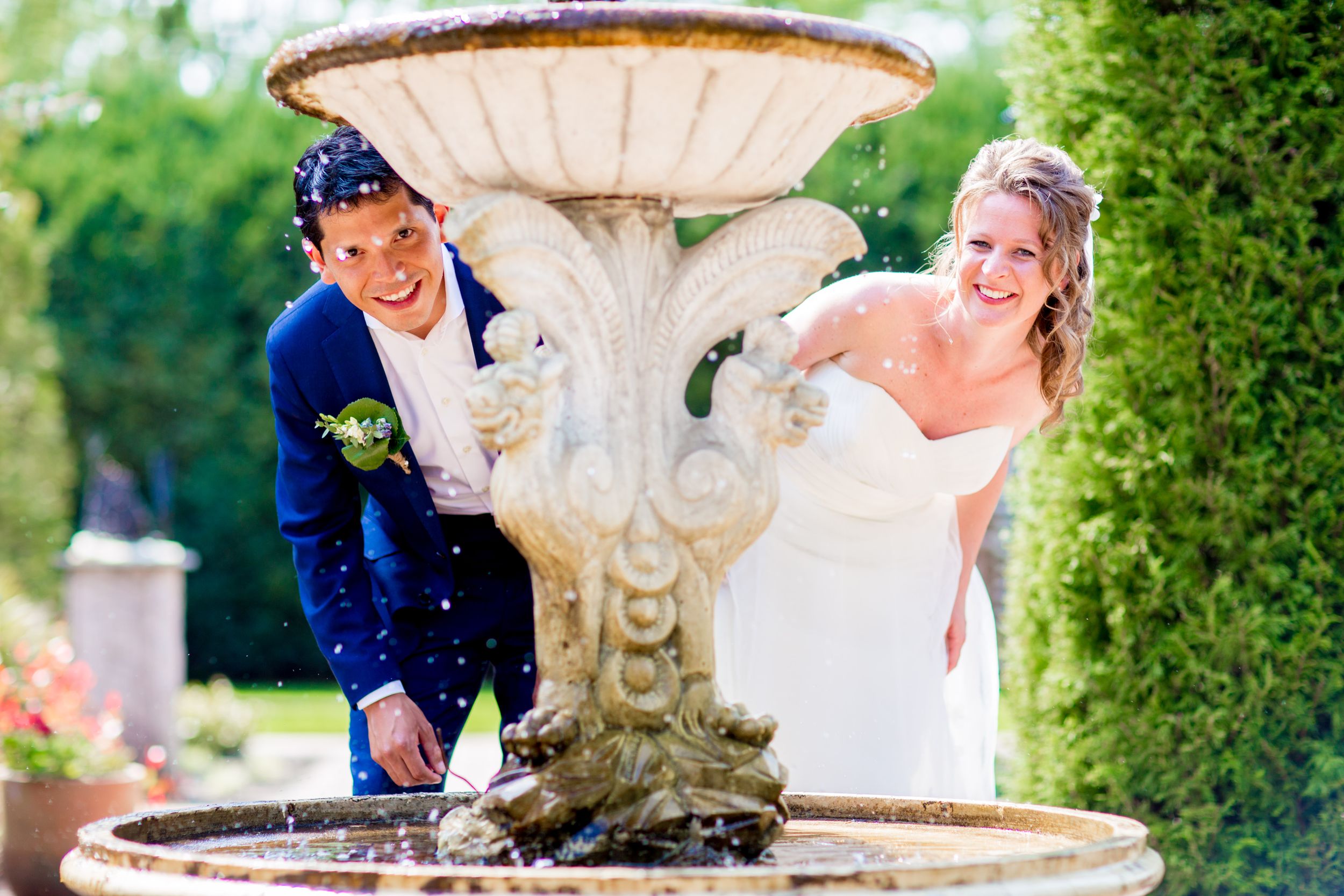Wedding couple laughing while posing by a decorative water fountain in garden setting.