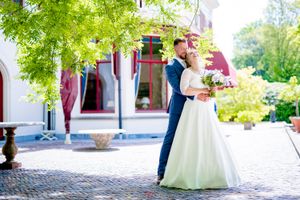 A couple in formal attire embraces on a brick patio outside an elegant historic building with white columns.