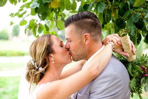 A romantic couple shares a tender kiss under green leaves on their wedding day.