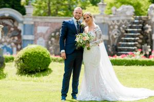 A couple in wedding attire walks hand in hand through a lush green garden on a sunny summer day.