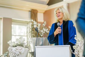 Speaker in a bright blue blazer presents at a formal event with white decorative accents in the background.