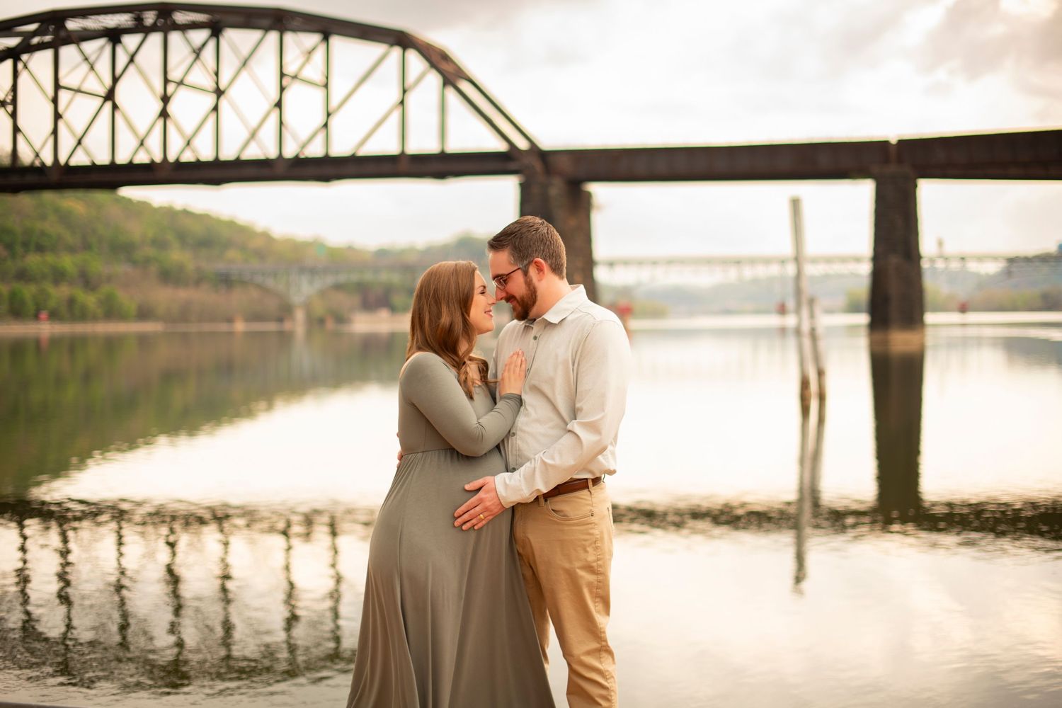 A couple shares an intimate moment by a lake with a steel bridge in the background during golden hour.