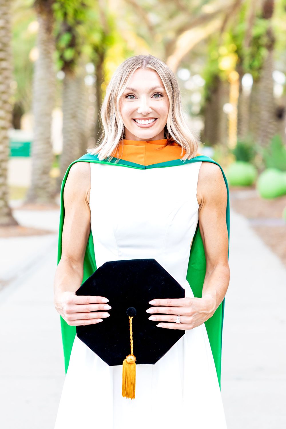 Smiling blonde woman in white dress holding black graduation cap wearing green and gold commencement regalia outdoors.
