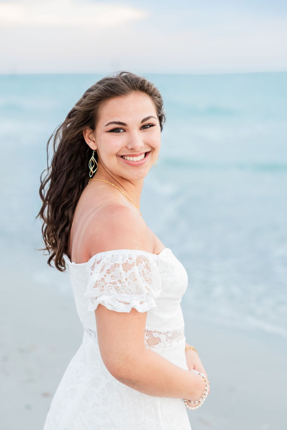 Young woman in white off-shoulder lace dress smiling at camera on a beach with turquoise water.