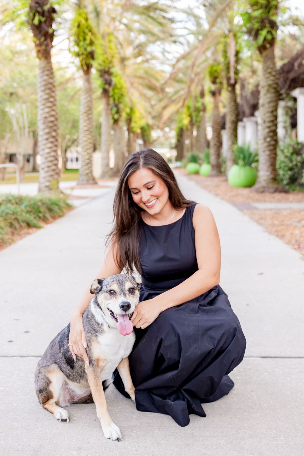 Smiling brunette woman in black dress crouches beside her happy dog on palm-lined path.