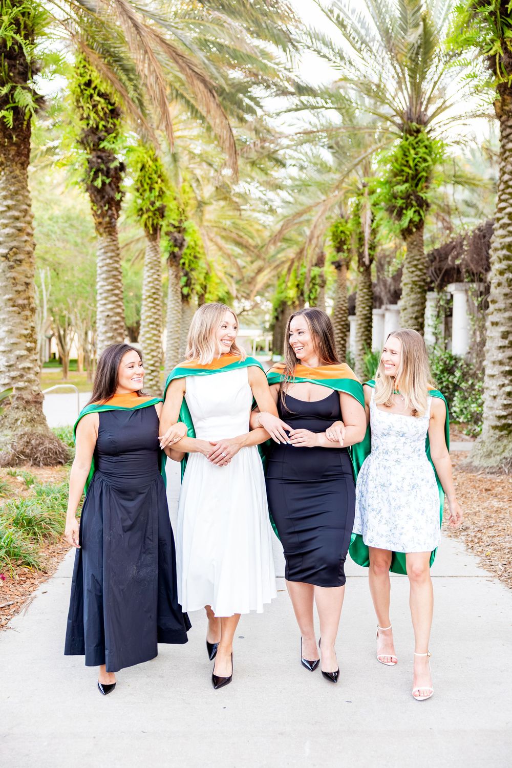 Four women in graduation hoods walk and laugh together on sunny palm-lined campus path.