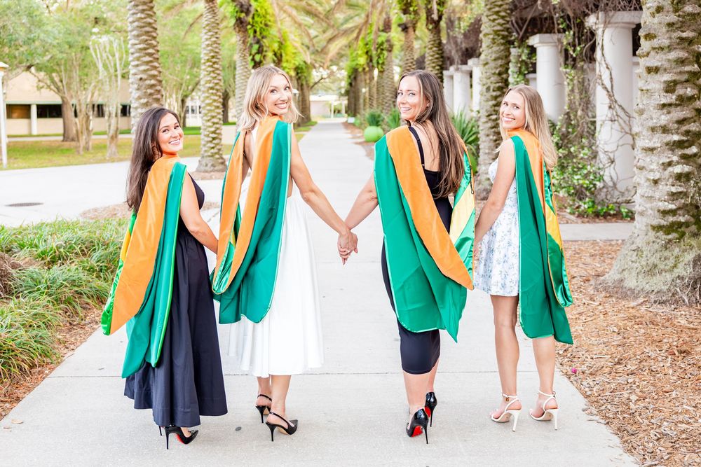 Four women in green and gold graduation hoods walk away holding hands on campus path.