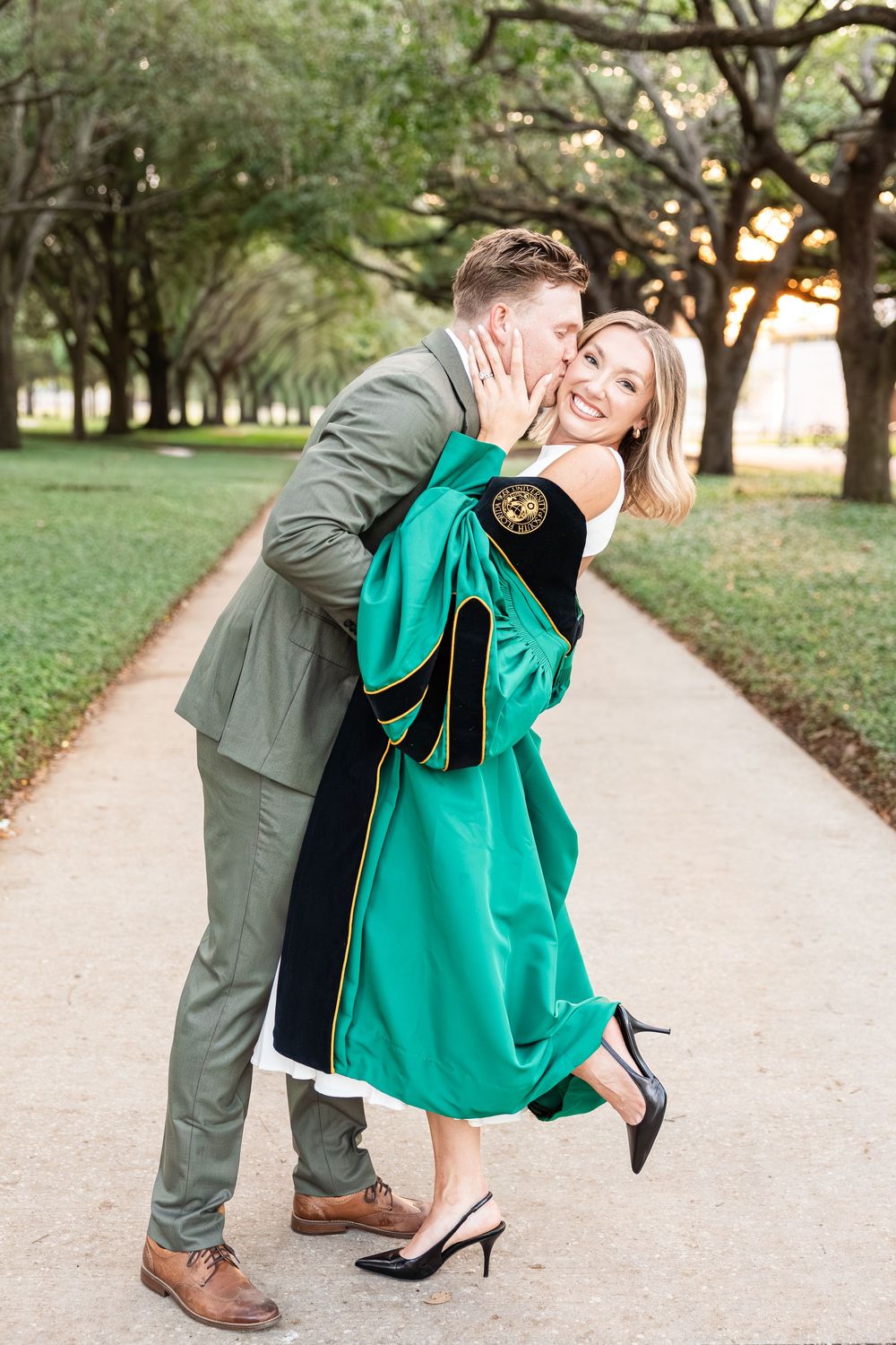 Man in gray suit dips and kisses blonde woman in green graduation gown on tree-lined path.