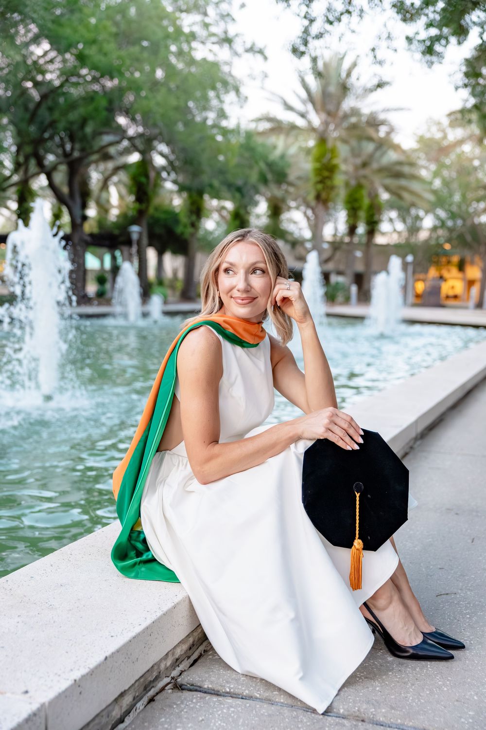 Blonde graduate in white dress and green hood sits by fountain holding black cap.
