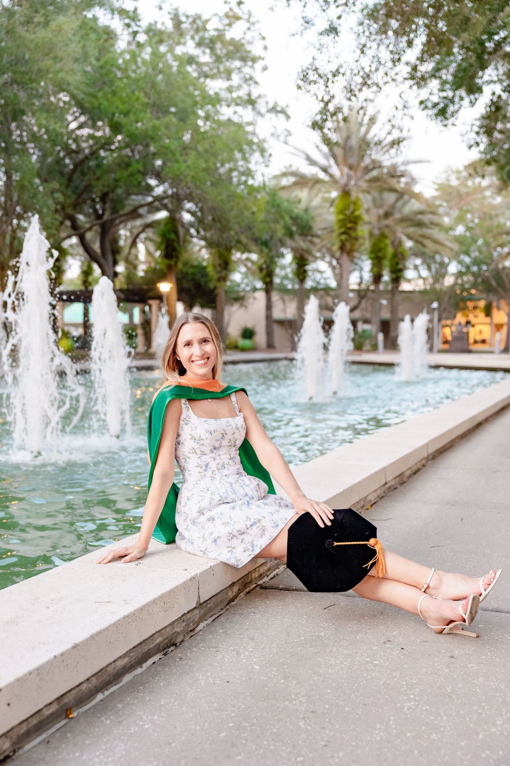 Smiling female graduate in floral dress with green hood sitting by a fountain holding cap.