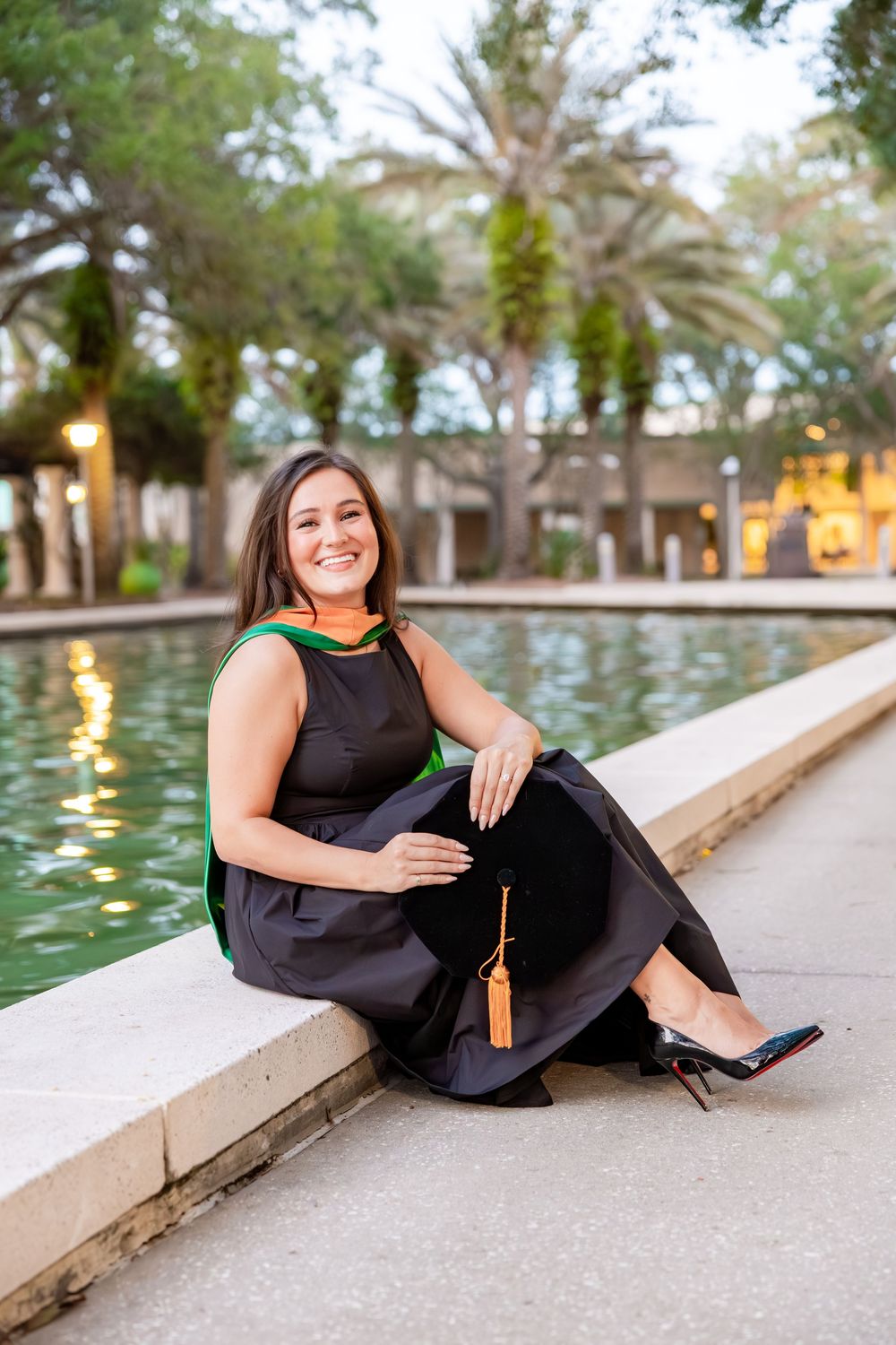 Brunette female graduate in black gown sitting by a reflective pond holding graduation cap.