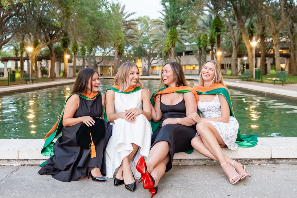 Four female graduates in black gowns with green and orange hoods laughing by a fountain.