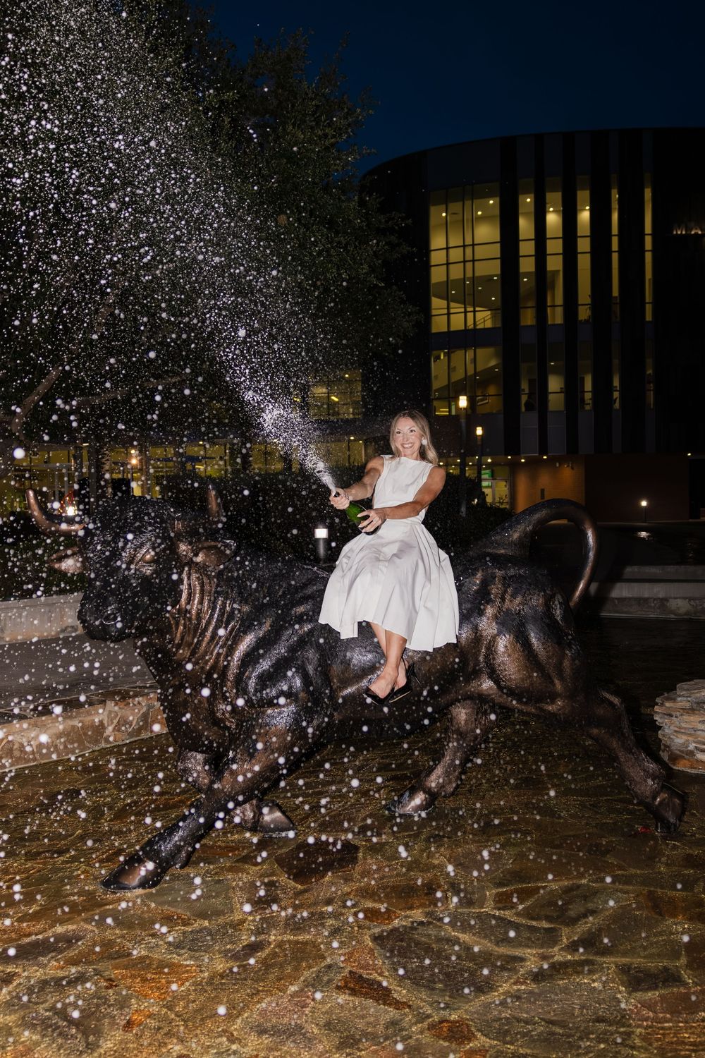 Woman in white dress riding a bronze bull statue at night with champagne spray.