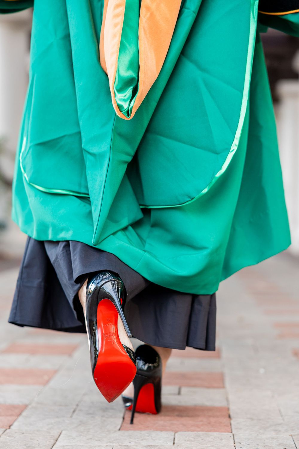 Close-up of graduation gown with red-soled Louboutin heels on brick pathway.