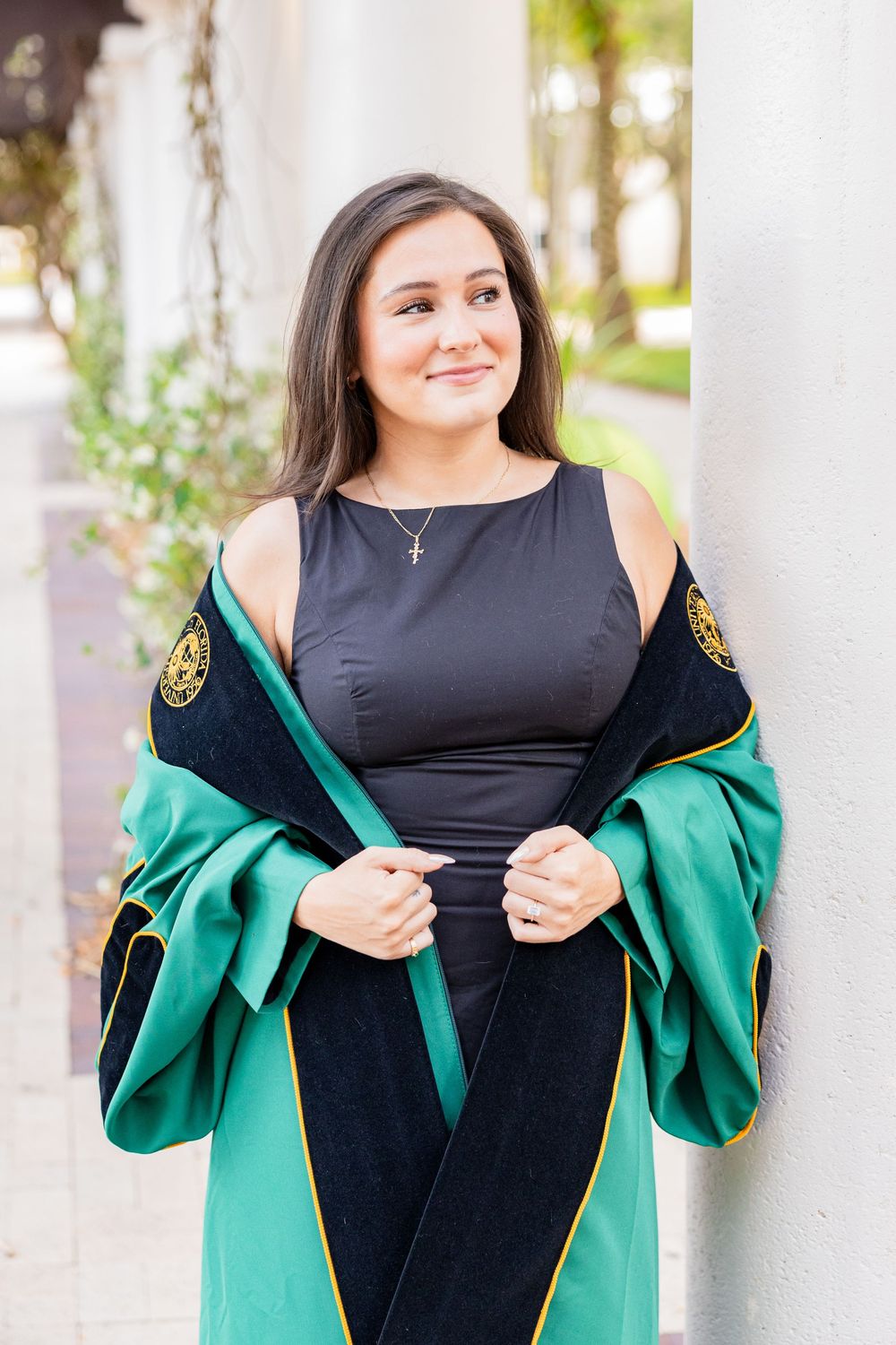 Brunette woman in black dress wearing green and black doctoral graduation regalia leaning against white column.