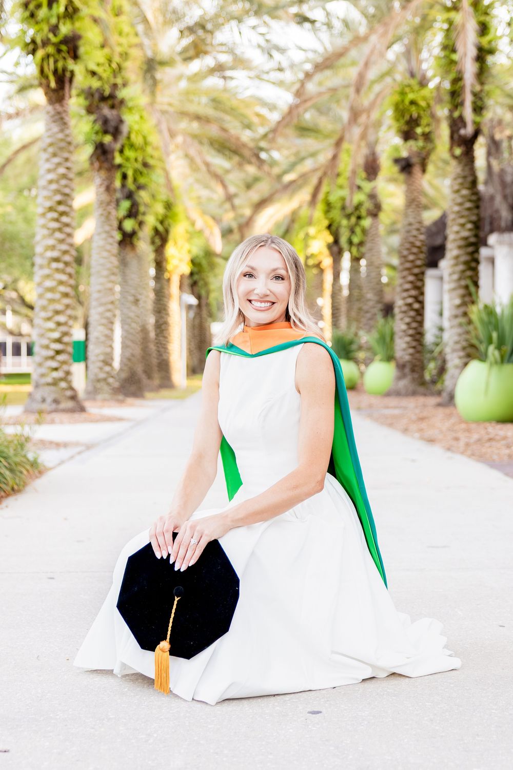 Smiling blonde graduate in white dress and green hood kneels with cap on palm-lined path.