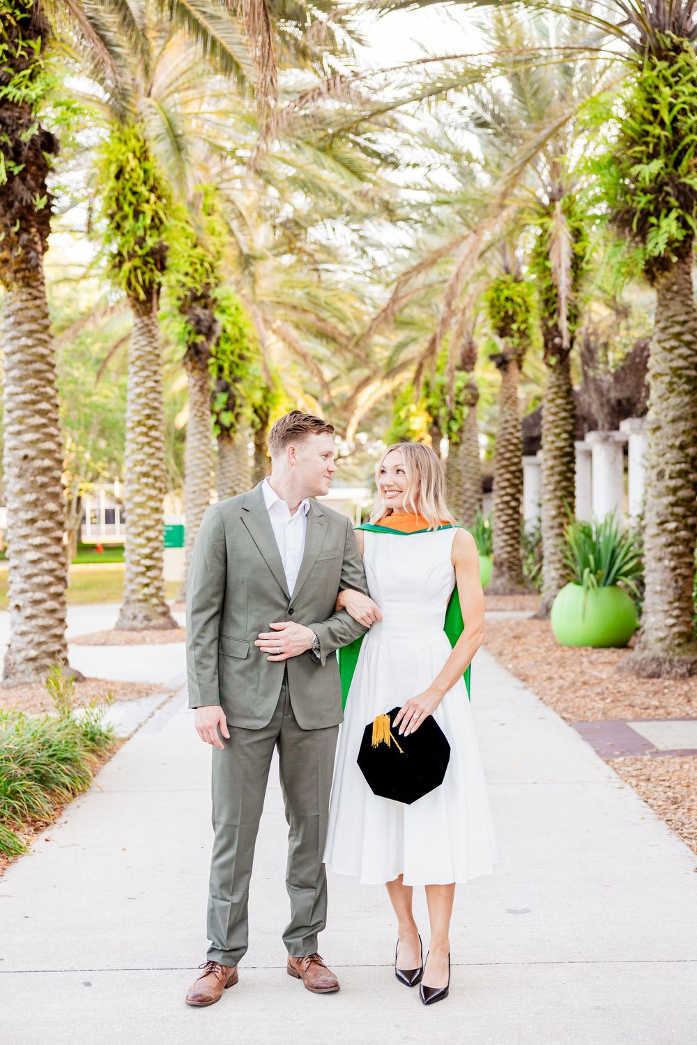 Graduate in white dress and green hood walks with man in gray suit among palm trees.