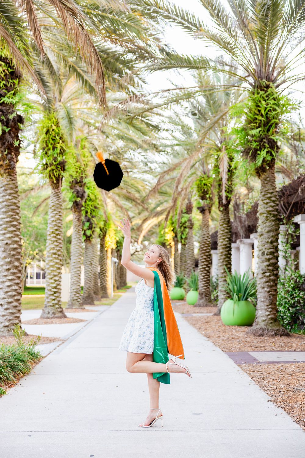 Blonde graduate tosses cap in air wearing white dress and green hood among palm trees.