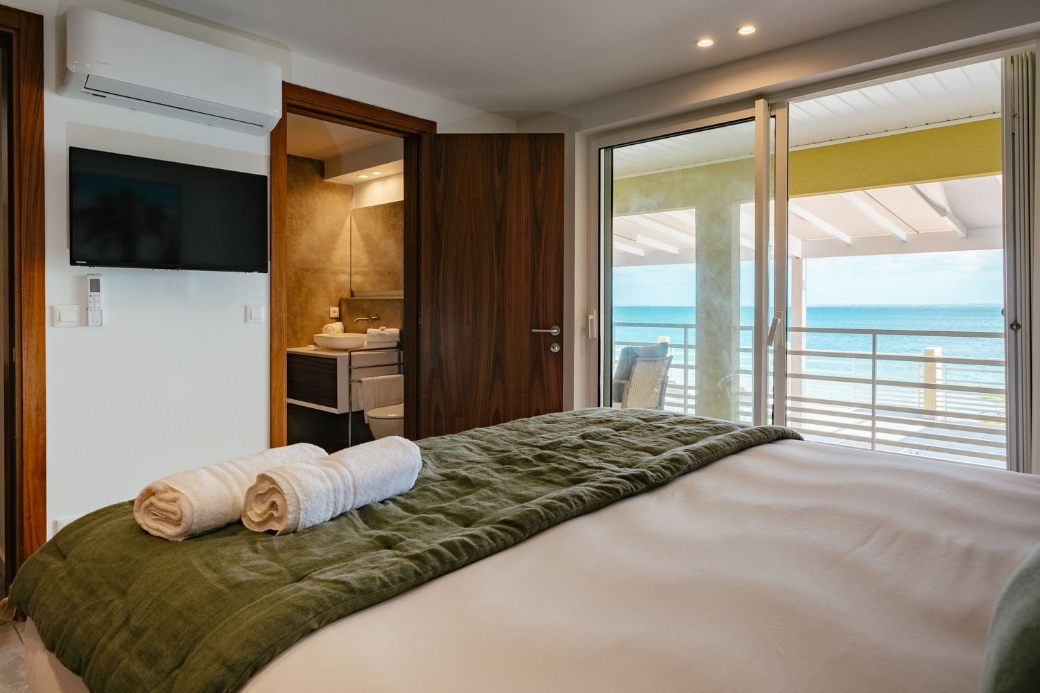 Modern hotel room interior with ocean view balcony, king bed with olive bedspread, and wall-mounted TV.