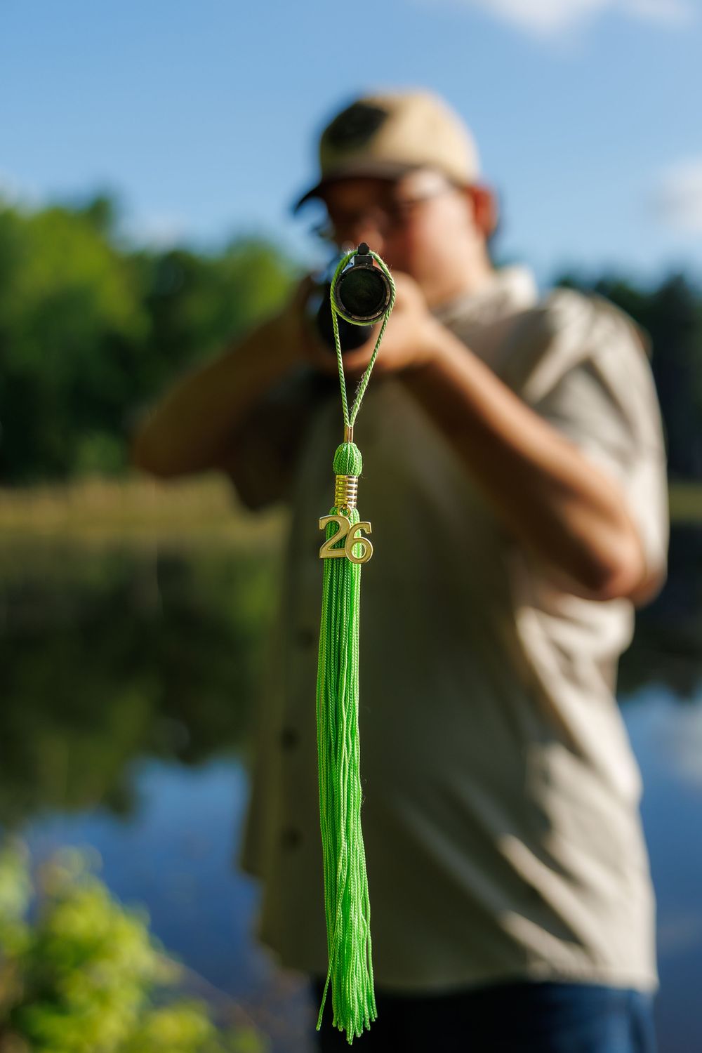 A green tassel charm with gold number 26 hangs in focus as a man aims a rifle near a lake.