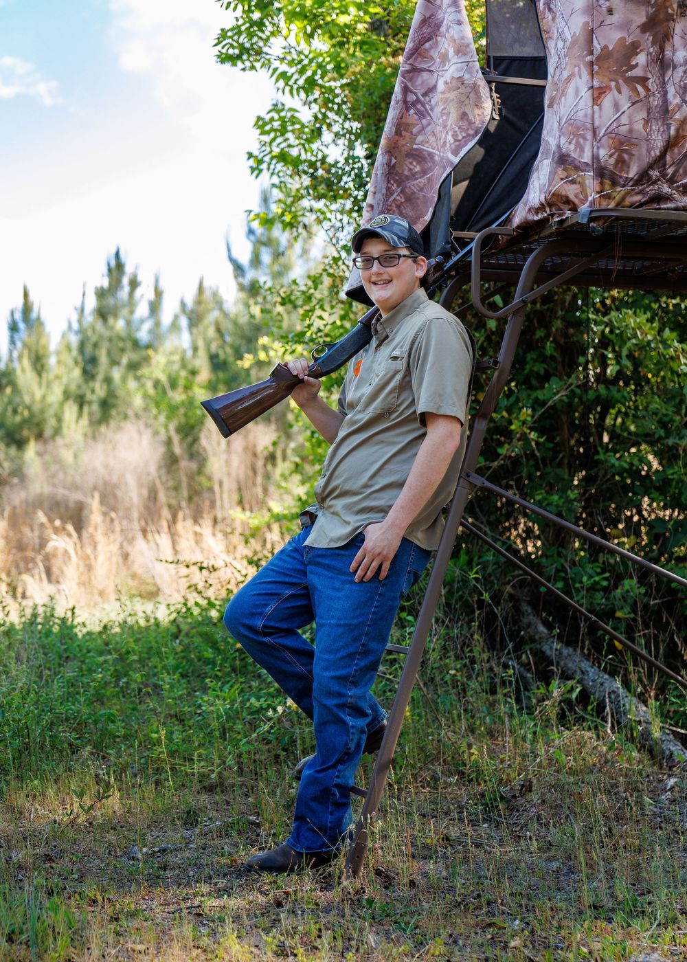 Young man in cap and glasses holds rifle while leaning against a hunting stand outdoors.