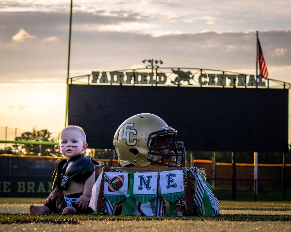 Baby in sports gear exploring football field at sunset with stadium in background.