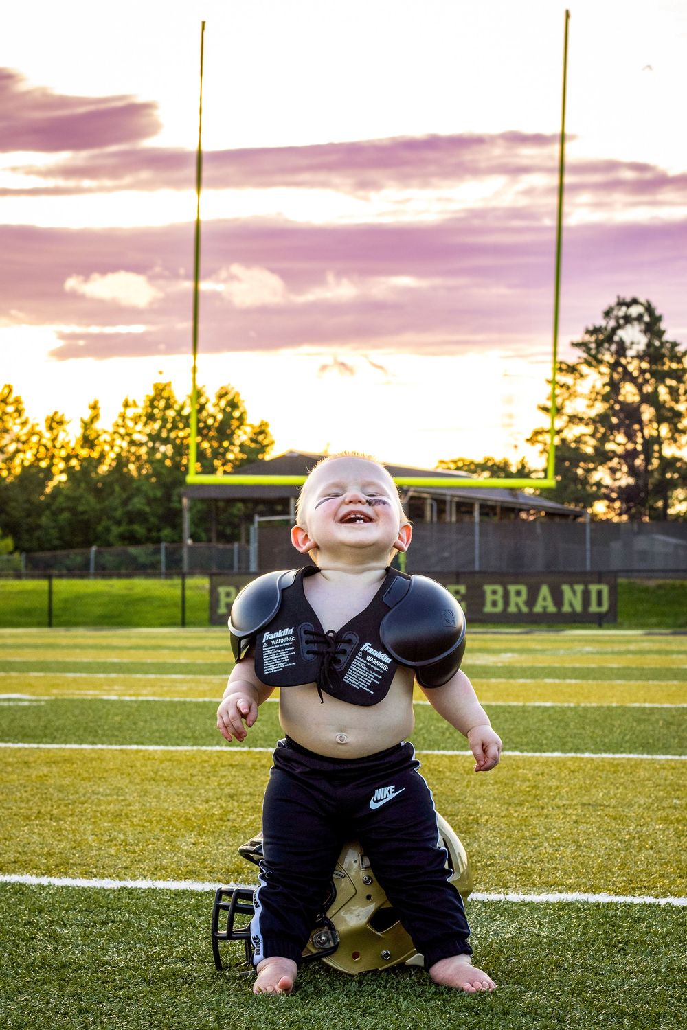 Small one year old wearing football shoulder pads kneels on a sports field at sunset with a joyful expression.