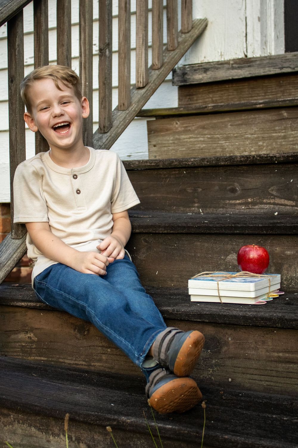 A cheerful boy sits on wooden steps with books and a red apple, during back to school mini session captured by Raley and Roots Photography.