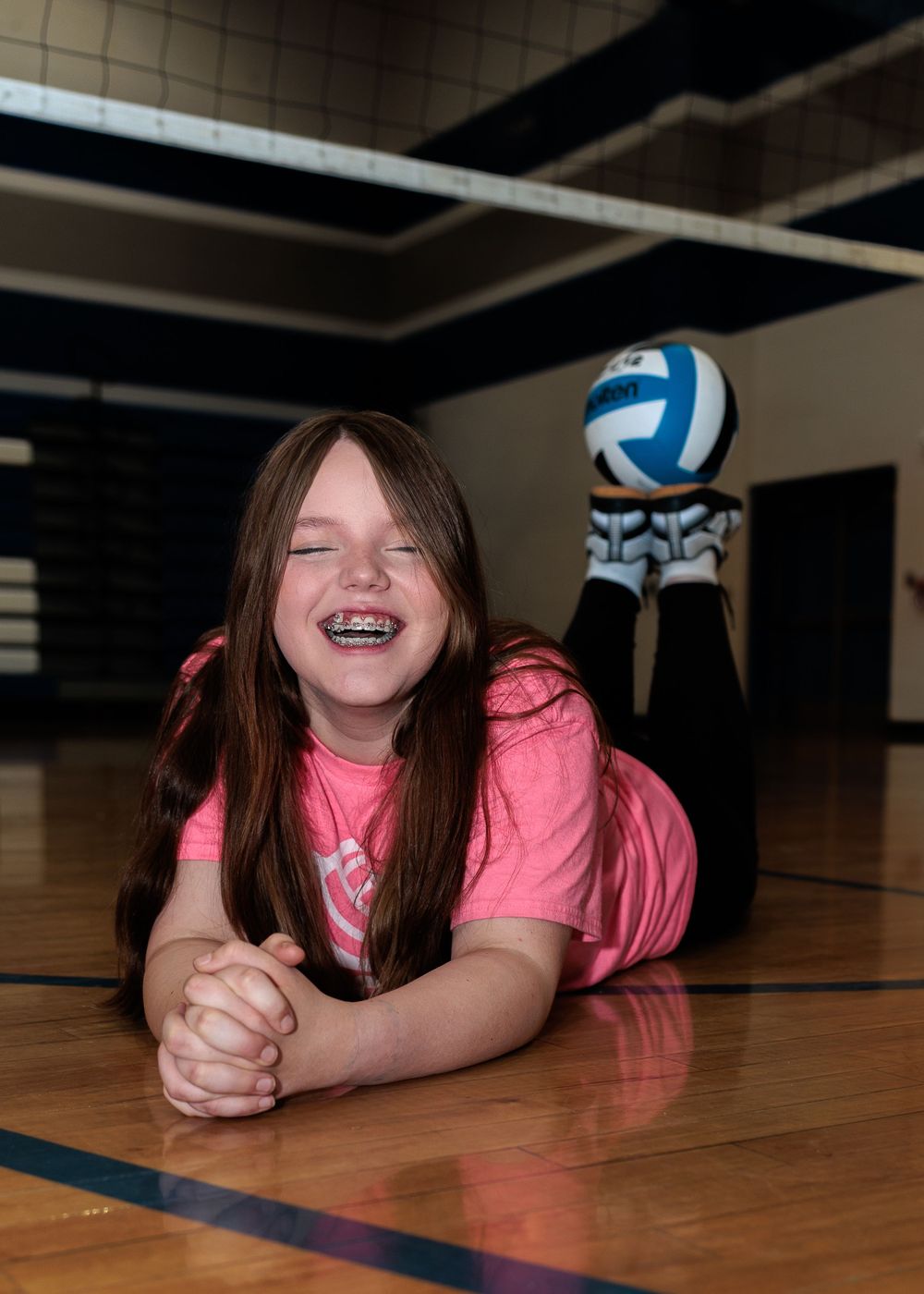 Young girl with braces laughing while lying on a gym floor with a volleyball behind her.