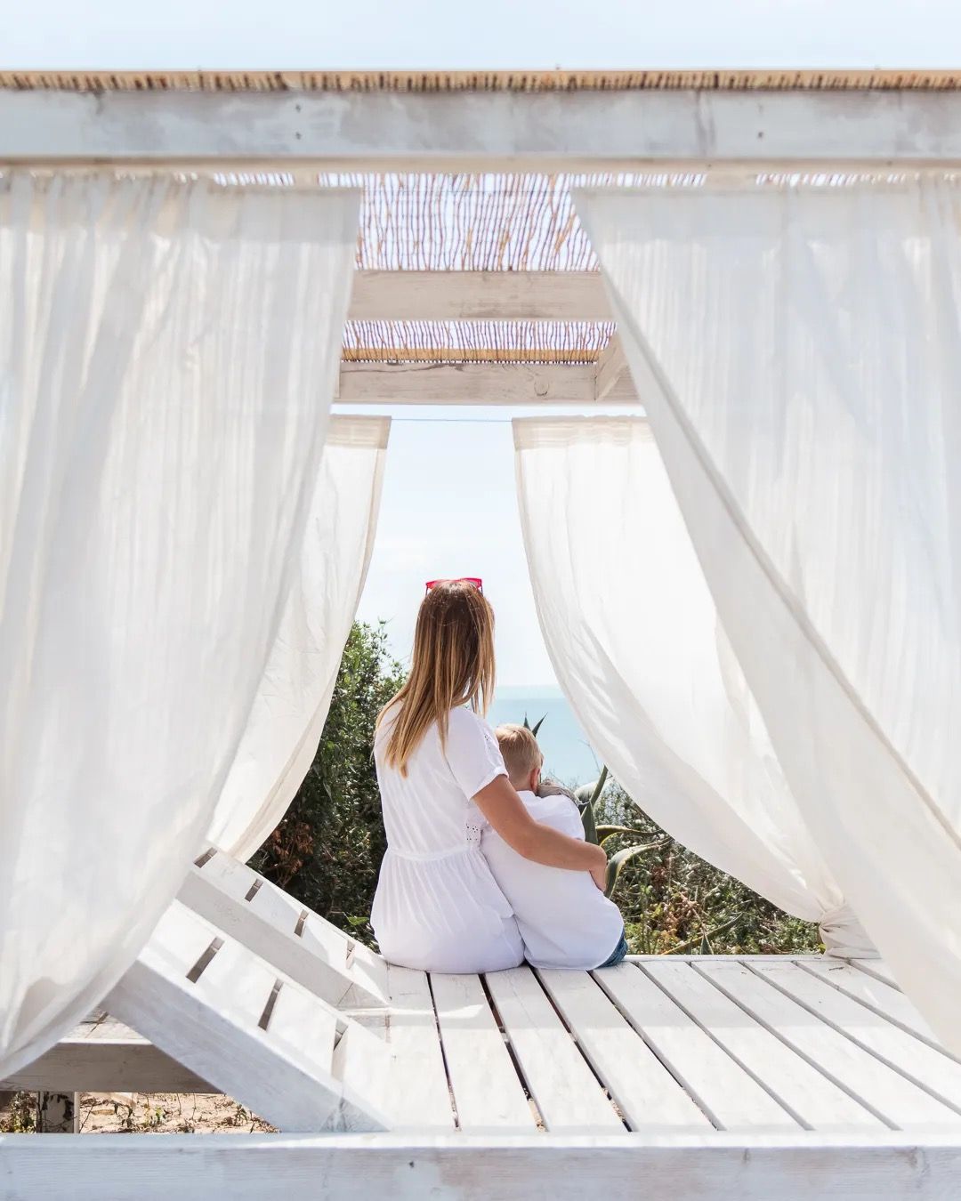 Girl relaxing with her son on a white hammock under soft curtains on a bright coastal-style porch.